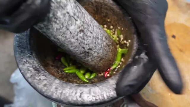 A high-angle, close-up shot of a person in black gloves using a traditional granite mortar and pestle to crush vibrant red and green chili peppers. This authentic process captures the preparation of a