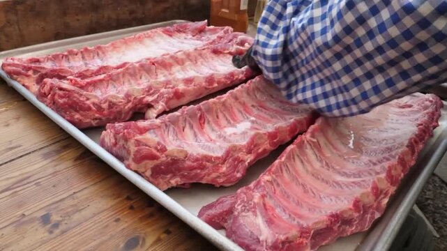 A detailed close-up shot capturing the process of preparing multiple racks of fresh, raw pork spare ribs. A hand is seen pouring a clear liquid binder from a bottle onto the meat, which is neatly arra