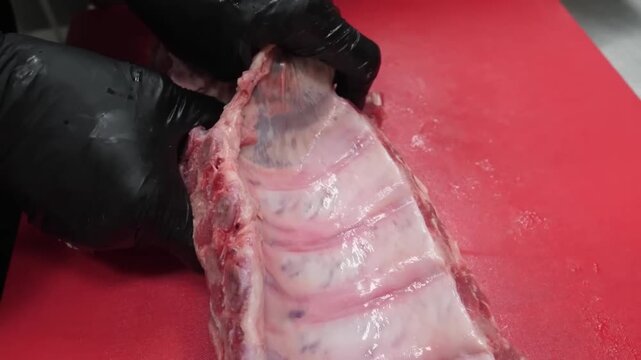 Close up of a chef in black latex gloves carefully removing the silver skin membrane from a fresh rack of pork baby back ribs on a red commercial cutting board in a restaurant kitchen. High-quality fo
