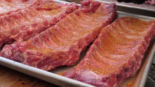 High-angle close-up footage of multiple racks of fresh, raw pork baby back ribs heavily coated with a savory red barbecue dry rub seasoning, resting on a professional stainless steel baking sheet in a