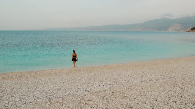 Woman facing calm turquoise sea on quiet Kefalonia pebble beach in summer morning