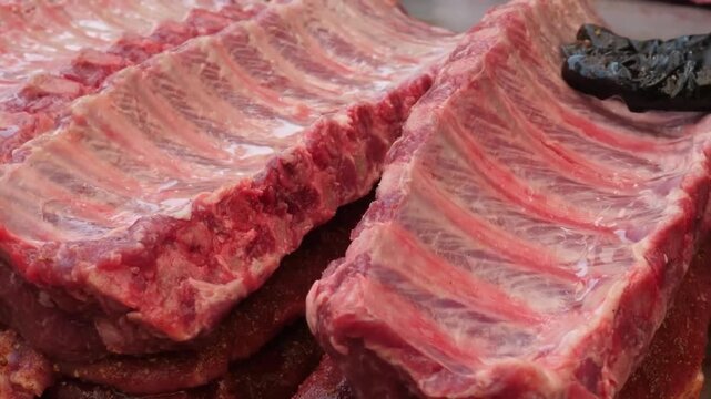 High-resolution footage showcasing multiple racks of raw pork spare ribs with visible bone structure and marbled meat texture. The ribs are arranged on a tray, ready for seasoning and slow-smoking at 