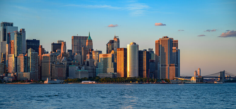 Lower Manhattan towers and Brooklyn Bridge at sunset. Warm light reflects from office facades beyond rippled water beneath scattered clouds.