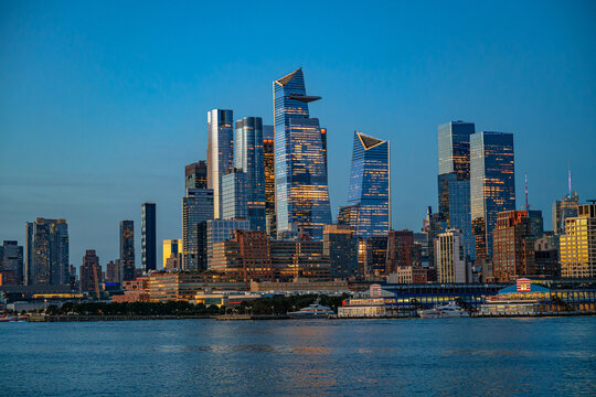 Hudson Yards skyscrapers reflected in evening river light. Layered office towers and waterfront buildings stand above boats on rippled water under a blue sky.