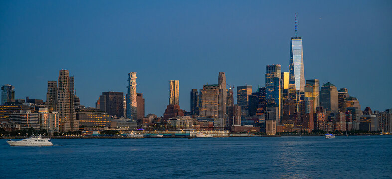 Lower Manhattan skyline with One World Trade Center. Wide river view shows illuminated towers, waterfront piers and a white yacht beneath a deep blue sky.
