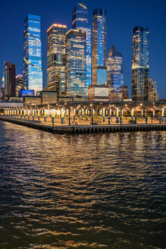 Hudson Yards skyline above Hudson River at blue hour. Illuminated Manhattan towers rise behind a covered waterfront pier with reflections on rippled water.