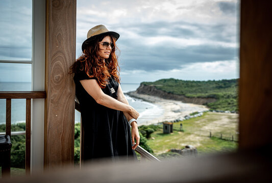 Woman rests beside porch column overlooking Long Island coast. Waist length portrait with folded arms, black dress, red hair, ocean cove, and grassy field in background.