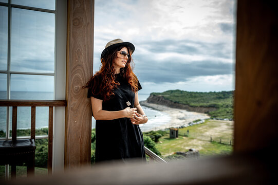 Coastal portrait of woman on wooden veranda in New York. Medium view frames a woman in sunglasses and straw hat against a beach, green cliffs, and gray clouds.