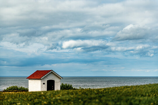White coastal shed under layered clouds on Long Island. Low composition places a red roofed structure behind blurred grass with ocean and broad cloud cover.