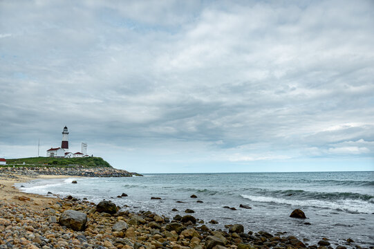 Montauk Point Lighthouse across rocky Atlantic shoreline. Wide coastal view places the distant tower beside open water, scattered rocks, waves, and layered clouds.