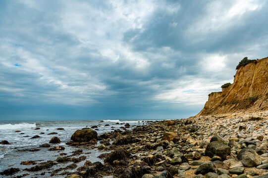 Eroded coastal bluff beside rocky Montauk shoreline. Brown cliff rises above boulders, seaweed, surf, and gray ocean under textured storm clouds.