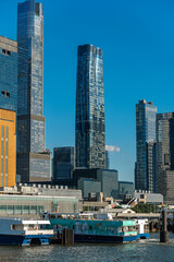 Fototapeta premium Passenger ferries below Hudson Yards residential towers, Manhattan. Vertical view frames glass high rises, terminal structures, dock pilings, and vessels along the river.