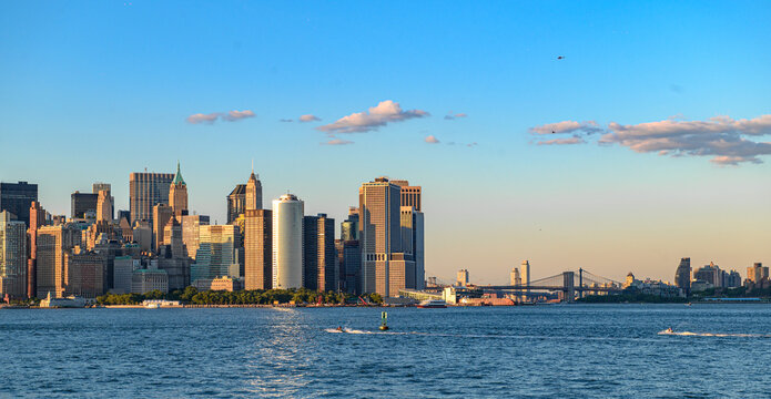 Lower Manhattan skyline and Brooklyn Bridge at sunset. Warm light reaches downtown towers as boats cross New York Harbor beneath helicopters and pink clouds.