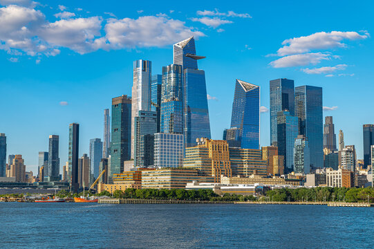 Hudson Yards skyline across Hudson River, Manhattan. Glass towers including Edge rise above mixed waterfront buildings beneath scattered white clouds.