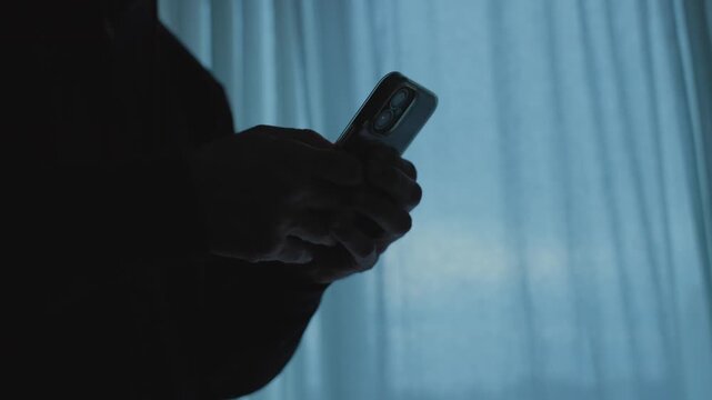 Scrolling a smartphone near a window at night in dark room in office or house. Man in dark clothes sending messages with mobile phone. Criminal activity by hacker or spy secretly communicating online