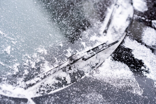 Close-up of a car windshield lightly covered with snow and frost. Two wiper blades rest against the glass, partially buried under thin layers of ice.