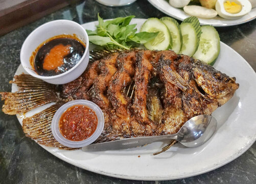 Crispy fried whole gourami fish served with sambal chili sauce sweet soy dip and fresh cucumber basil. Traditional Indonesian dish with authentic street food presentation.