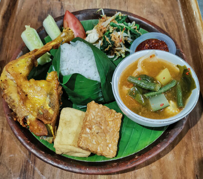 Authentic Indonesian chicken rice meal served with vegetable soup, tofu, tempeh, urap vegetables, sambal, cucumber and tomato on banana leaf over a rustic clay plate.