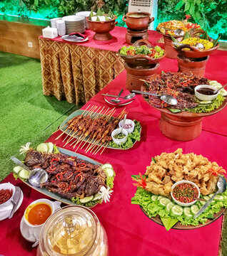 Indonesian buffet catering table with satay grilled meat fried chicken sambal and traditional dishes served in clay pots. Rich food spread for events and celebrations.