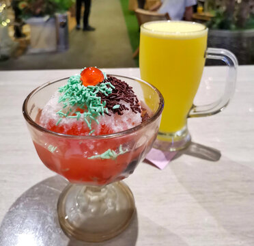 Authentic Indonesian Es Campur featuring shaved ice, colorful sprinkles, cherry, sweet syrup and jelly served with a refreshing yellow fruit beverage in a cozy restaurant.