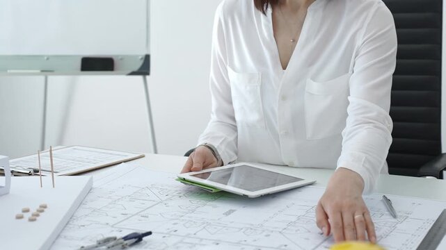 Woman architect analyzing blueprints and architectural models with a digital tablet on a clean desk, working on a construction project in a bright office environment