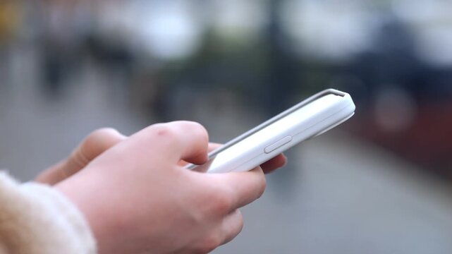 Close-up of hands holding a smartphone outdoors. Person wears fuzzy sleeve, thumbs scrolling. Focus on device, background blurred. Suitable for social media, tech, or communication themes