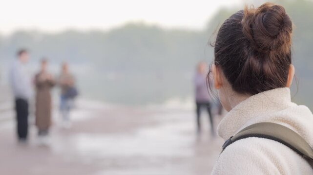 Woman with backpack walks forward on path, focused ahead. Blurred figures behind suggest movement and distance. She moves calmly, purposeful step toward open space. Ideal for travel, urban exploration
