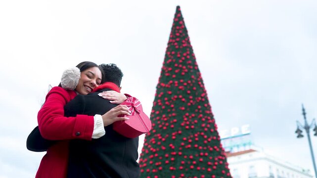 Romantic young couple hugging and exchanging Christmas gifts at Puerta del Sol square in Madrid, Spain, with a large illuminated Christmas tree in the background during the festive season