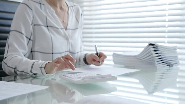 Woman's hands using a calculator and holding a pen, working with financial documents and a pile of papers on a clear desk in an office setup