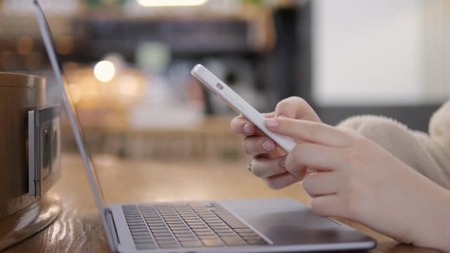 Person uses smartphone while laptop sits open nearby. Focus on hands holding phone. Background blurred cafe setting. Neutral expression, calm and focused. Ideal for digital communication, remote work