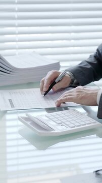 Financial professional analyzing data and calculating expenses using a desktop calculator, surrounded by stacks of documents, a pen, and eyeglasses on a glass desk