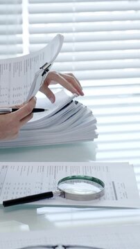 Person examining business documents on a desk with a magnifying glass, analyzing financial reports for an audit, tax, or accounting process