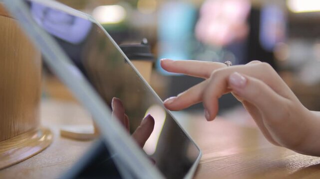 Close-up of hand scrolling digital tablet on wooden table. Fingers tap screen with focus, motion blur suggests active use. Soft bokeh background implies relaxed, indoor setting. Person appears engaged