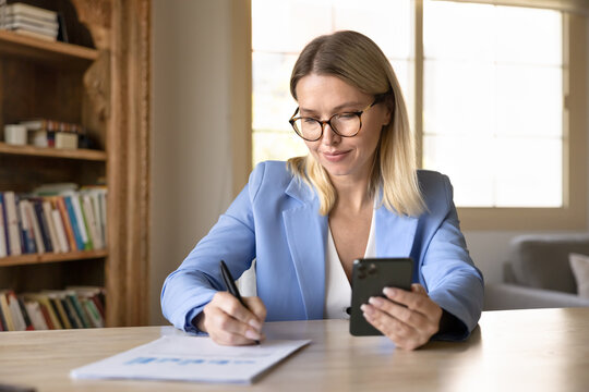Businesswoman sitting at workplace desk with smartphone, browsing, writing notes in report documents displaying diagrams, making financial data analysis, researching information, planning strategy