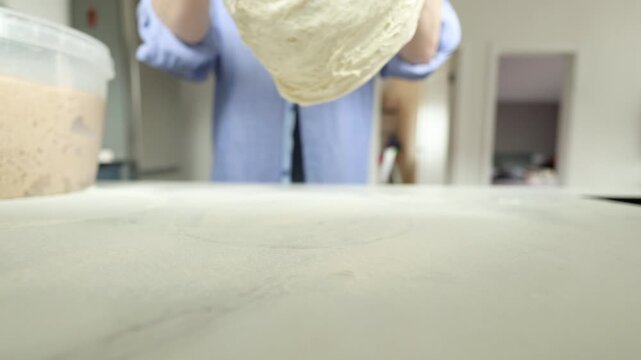 Proofed dough is lifted by blurred hands over pale kitchen surface beside clear container, with soft daylight, shallow focus, calm home setting, and tactile bread-making process visible throughout