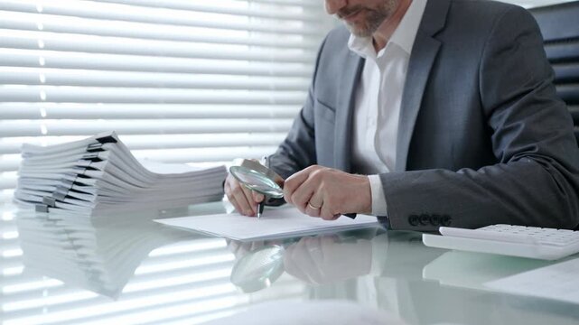 Business professional meticulously inspecting a contract or official document with a magnifying glass and pen while working at a desk with a stack of paperwork