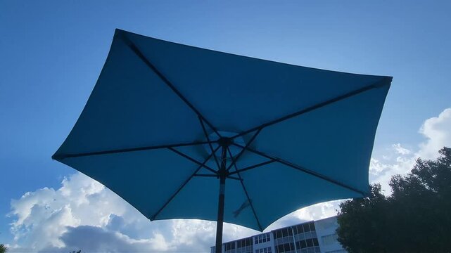 Cinematic outdoor video featuring a large blue patio umbrella silhouetted against bright sunlight, dramatic clouds, and a tropical sky. Ideal for vacation, resort, leisure, summer relaxation, hospital