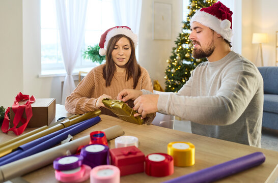 Christmas couple in Santa hats wrapping surprise gift box with ribbon and gold paper. Man and woman siting at table together to tie bow from rope, preparing festive packaging in living room at home