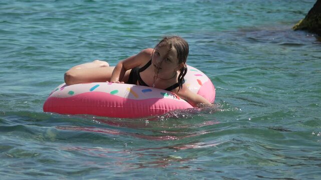 Girl swimming pool, child playing on inflatable donut float in clear sea water during summer vacation at tropical beach on sunny day