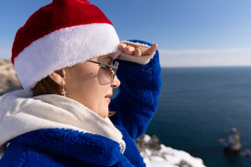 Young woman wearing santa hat looks into distance standing by blue sea during sunny winter day on coastal cliff side © svetograph