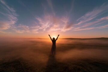 Fototapeta premium Silhouette of a woman with arms raised in a misty sunrise landscape, featuring a glowing atmospheric sky and vast copy space for wellness and inspiration