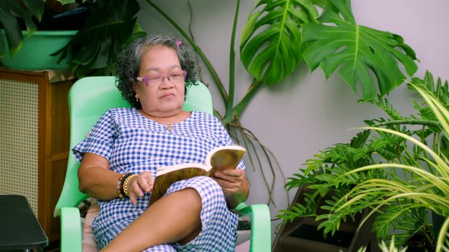 Senior Asian woman relaxing and reading a book in a lush green home garden, elderly retirement lifestyle concept.