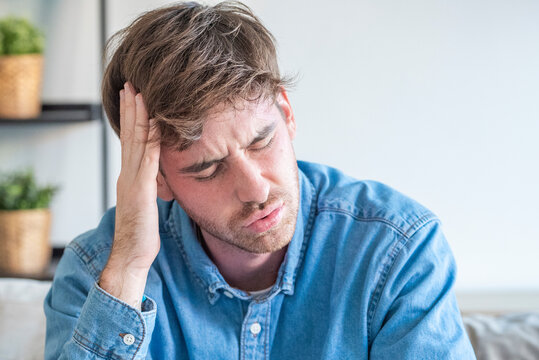 Young man holding his head in pain sitting on a couch, stressed and suffering from a headache or migraine at home, feeling tired and anxious