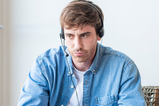Man wearing headset looking thoughtful during a remote customer service call in casual denim shirt, working from home as a call center agent showing concentration and mild frustration