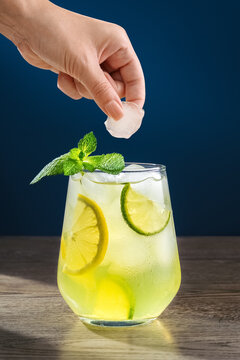 A hand drops an ice cube into a glass of cocktail. Lemon slices and a mint leaf float in the refreshing drink. Bright yellow color in a glass on a blue background. Vertical photo