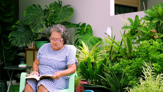 Senior Asian woman relaxing and reading a book in a lush green home garden, elderly retirement lifestyle concept.