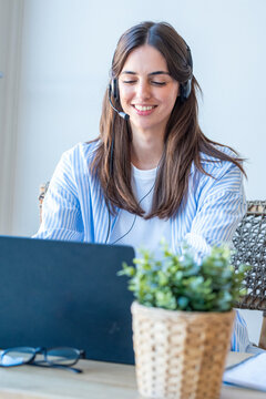 Young woman wearing a headset smiling while working on a laptop at home, remote customer service or online meeting in a bright home office setting