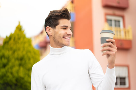 Young caucasian man holding a take away coffee at outdoors with happy expression