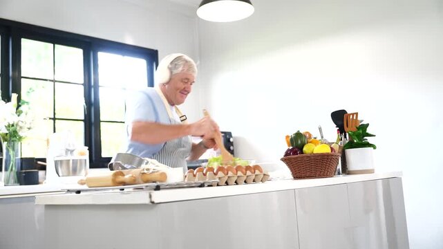 An elderly grandfather, happily wearing headphones, dances to himself while standing on the kitchen counter, holding a salad stick, making a salad for breakfast in his home kitchen.
