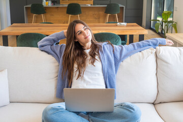 Young woman relaxing on a sofa with a laptop, stretching and smiling while working from home in a bright modern living room, feeling comfortable and content.